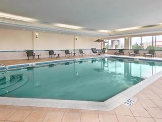 An indoor hotel swimming pool with clear blue water, surrounded by lounge chairs and large windows allowing natural light.